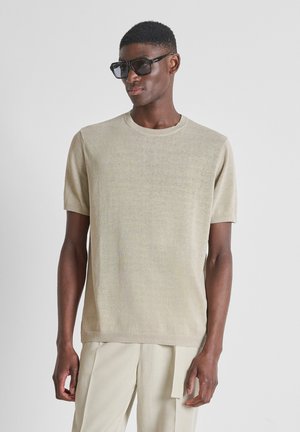Jonge man met beige gebreide korte mouw shirt, bijpassende beige broek met ceintuur detail en grote zwarte vierkante zonnebril, zijwaarts kijkend.