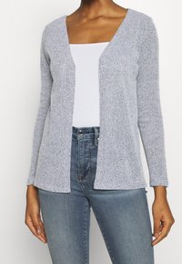 Kobieta w jasnoszarej, strukturalnej kardiganie, założonej na białą, prążkowaną bluzkę i wysokie niebieskie dżinsy, stojąca na tle gładkiej, białej ściany.