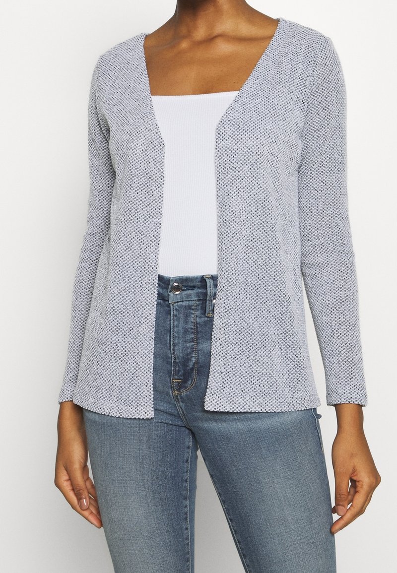 Kobieta w jasnoszarej, strukturalnej kardiganie, założonej na białą, prążkowaną bluzkę i wysokie niebieskie dżinsy, stojąca na tle gładkiej, białej ściany.