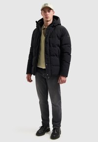Zwarte geïsoleerde pufferjas met een capuchon, voorzien van een ritsluiting en twee voorkockets, gedragen over een beige overhemd met knopen en donkere jeans.