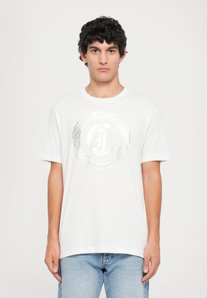 Young man with curly hair stands facing forward wearing a white Just Cavalli T-shirt with a silver snake logo and light blue jeans.
