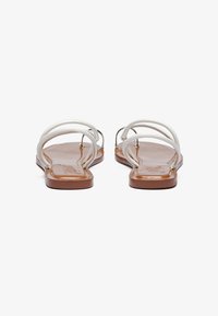 Hvide flade sandaler med to tynde stropper med struktureret finish, sat på en brun flad sål med en glat overflade.