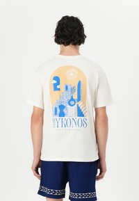 Witte t-shirt met korte mouwen en een blauw en geel grafisch ontwerp met trappen, planten en de tekst "MYKONOS PURE PARADISE."