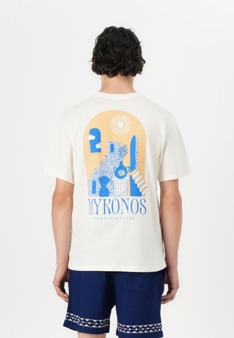 Weißes T-Shirt mit kurzen Ärmeln und einem blauen und gelben grafischen Design, das Treppen, Pflanzen und den Text "MYKONOS PURE PARADISE" zeigt.