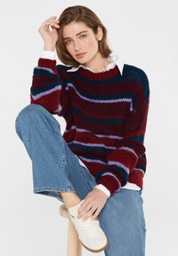 Gebogener burgunderfarbener Pullover mit blauen und lila Streifen, lockerer Schnitt, gerippte Bündchen und Saum. Kombiniert mit hellblauen Jeans und weißen Turnschuhen.
