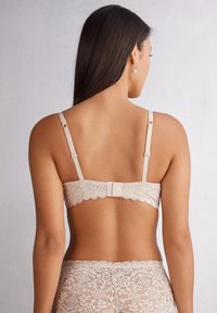 Sujetador con espalda de encaje en color nude con tirantes ajustables. Presenta bordes de encaje en forma de ondas y un cierre de gancho y ojo. Textura suave.