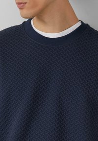 Granatowa teksturowana bluza z wzorem tkanym, prążkowanym okrągłym dekoltem oraz warstwową białą koszulką widoczną przy kołnierzu.