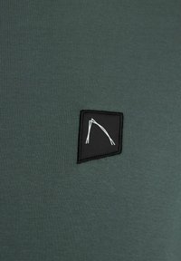 Dunkelgrüner Stoff mit gerippter Textur, der in der oberen Ecke ein schwarzes besticktes Logo-Patch mit einem minimalistischen weißen Design zeigt.