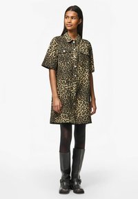 Abito camicia con stampa leopardata in beige e nero, dotato di maniche corte, tasche sul petto e chiusura a bottoni automatici. Abbinato a stivali neri al ginocchio.