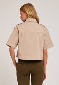 Giacca beige cropped con maniche corte, realizzata in un tessuto liscio, dotata di colletto a punta e dettaglio della cucitura sul retro.