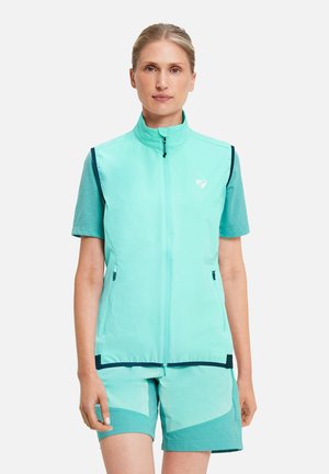 Frau trägt eine ärmellose türkise Weste mit Reißverschluss über einem passenden kurzärmligen Shirt und zweifarbigen türkisen Shorts, steht vor weißem Hintergrund.