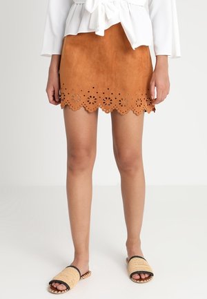 Femme portant une mini-jupe en daim beige avec un ourlet festonné à découpes florales, une blouse blanche et des sandales beige tissées à bout ouvert.