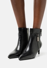 Botas de cuero negras de punta afilada hasta el tobillo con cremalleras laterales, llevadas sobre piernas desnudas contra un fondo blanco.