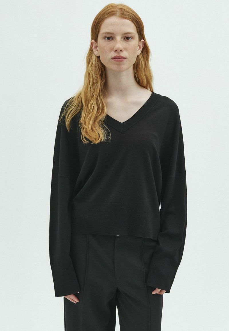 Jeune femme aux longs cheveux roux portant un pull noir ample à col en V et un pantalon noir, debout devant un fond clair uni.