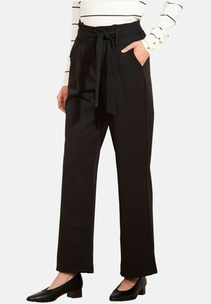 Woman wearing black high-waist wide-leg trousers with tied belt, one hand in pocket, paired with black low-heeled shoes and striped white top.