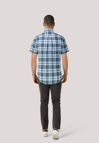 Kurzärmeliges Karomusterhemd in Blau, Grün und Weiß mit abgerundetem Saum. Kombiniert mit dunkelgrauen Hosen und weißen Sneakern.