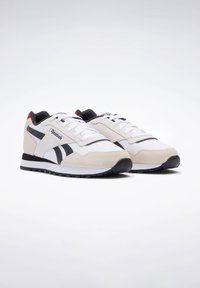 Weiße und beige Sneakers mit schwarzen Akzenten, aus Wildleder und synthetischen Materialien, mit einer Schnürsenkeldesign und einer Mesh-Zunge. Reebok-Logo sichtbar.