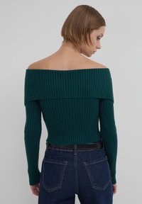 Maglione a spalle scoperte in verde scuro con maniche lunghe, design aderente e finitura testurizzata. Abbinato a jeans blu a vita alta.
