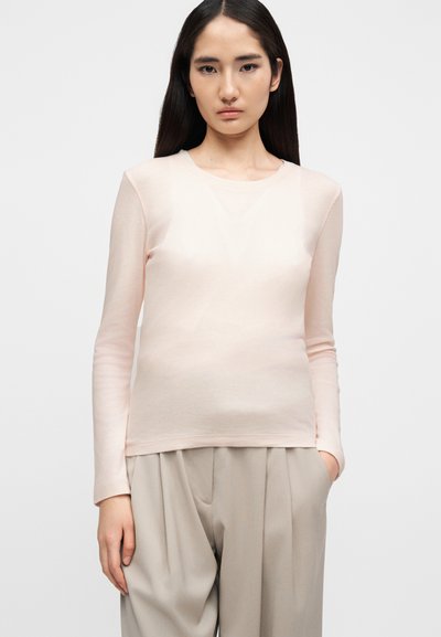 Ανοιχτό ροζ μπλουζάκι με μακριά μανίκια από μαλακό ύφασμα, με στρογγυλή λαιμόκοψη και στενή εφαρμογή, σε συνδυασμό με φαρδιά μπεζ παντελόνι.