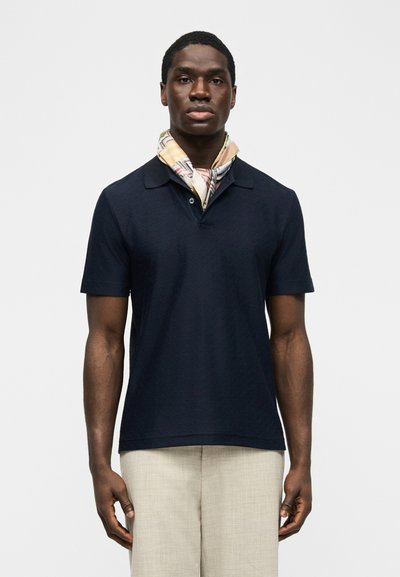 Άνδρας που φοράει μπλε κοντομάνικο πόλο, μπεζ παντελόνι και μεταξωτό μαντήλι με σχέδια δεμένο γύρω από τον λαιμό του, στέκεται μπροστά σε λευκό φόντο.