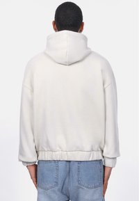 Sweat à capuche couleur crème avec une coupe décontractée, taille élastique et poignets côtelés. Tissu doux avec des détails de couture visibles.
