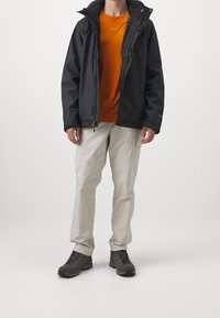 Chaqueta impermeable negra, camiseta naranja, pantalones cargo beiges y zapatos de senderismo oscuros. La chaqueta cuenta con capucha y cremallera frontal.