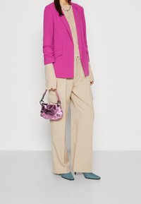 Fuchsia kavaj över en beige topp, i kombination med beige vida byxor. Håller en glänsande rosa väska; bär teal spetsiga skor. En enkel silverkedja synlig.