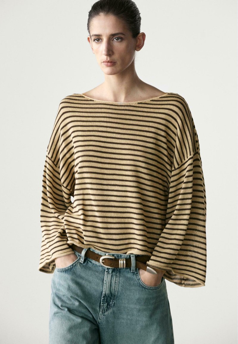 Femme aux cheveux foncés portant un pull ample beige à rayures noires, rentré dans un jean bleu clair avec une ceinture marron, debout les mains dans les poches.