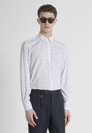 Witte blouse met een zwart geometrisch patroon, lange mouwen en een standaardkraag, gecombineerd met donkere broeken.