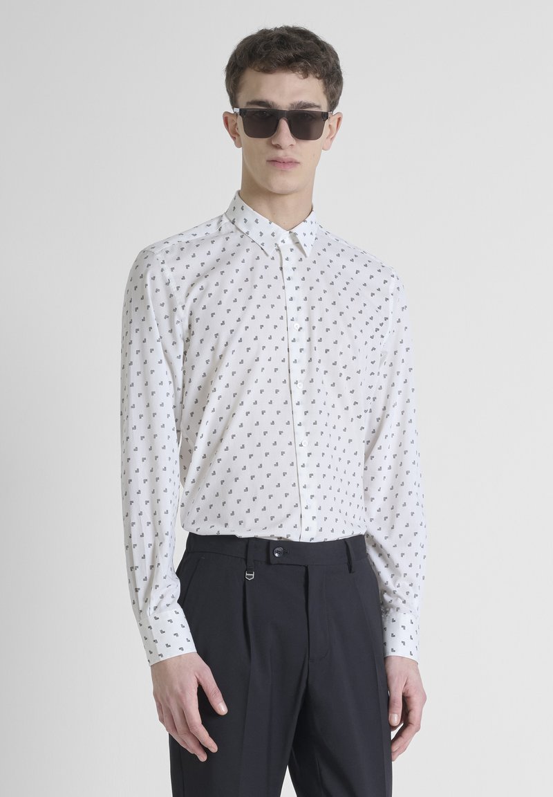 Camicia bianca con bottoni, caratterizzata da un motivo geometrico nero, maniche lunghe e colletto classico, abbinata a pantaloni scuri.