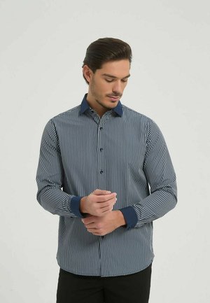 Homme ajustant les manches d'une chemise boutonnée à fines rayures bleu marine et blanche, avec col et poignets bleu foncé, debout devant un fond clair uni.