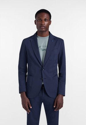 Mand iført en marineblå jakkesætjakke og bukser med en grøn grafisk t-shirt, stående mod en ensfarvet hvid baggrund.