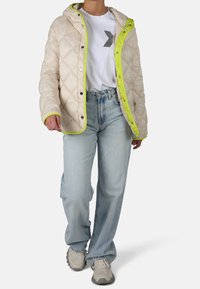 Giacca in crema trapuntata con rifiniture verde neon e cappuccio, abbinata a jeans azzurri chiari e sneakers beige. La giacca è dotata di bottoni a pressione.