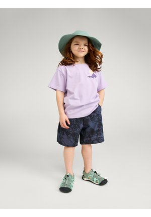 Young child with red hair wearing a green wide-brim hat, lavender T-shirt, dark patterned shorts, and green sandals, standing with hands in pockets.