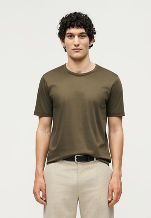 Jeune homme aux cheveux bouclés portant une chemise courte à manches courtes olive et un pantalon beige avec une ceinture noire, debout devant un fond uni clair.