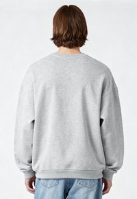 Personne aux cheveux bruns courts, portant un sweat-shirt gris clair uni et un jean bleu, debout de dos sur un fond uni.