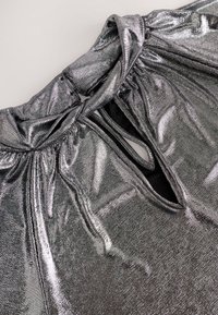 Tissu argenté métallisé avec une découpe en forme de trou de serrure et un col froncé montrant une texture détaillée et une surface brillante et réfléchissante.