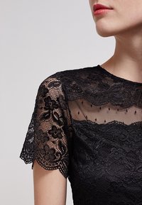 Černý krajkový top s květinovými vzory, průsvitnými detaily a krátkými rukávy. Hladká textura s kulatým výstřihem.