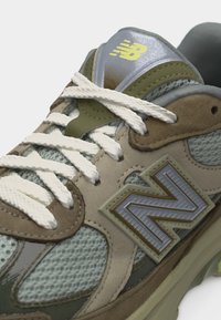 Αθλητικό παπούτσι New Balance σε μπεζ και ελιά πράσινο χρώμα με λευκά κορδόνια, πλέγμα, σουέντ επενδύσεις και μεγάλο λογότυπο "N" στο πλάι.