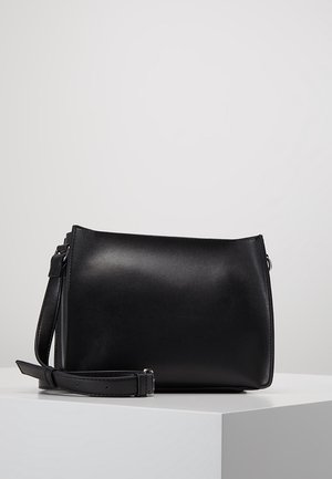 Schwarze rechteckige Leder-Crossbody-Tasche mit verstellbarem Riemen, die auf einem weißen Podest vor neutralem Hintergrund liegt.