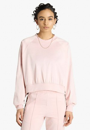 Donna che indossa una felpa a maniche lunghe rosa chiaro e pantaloni coordinati, in piedi frontalmente su uno sfondo bianco semplice.