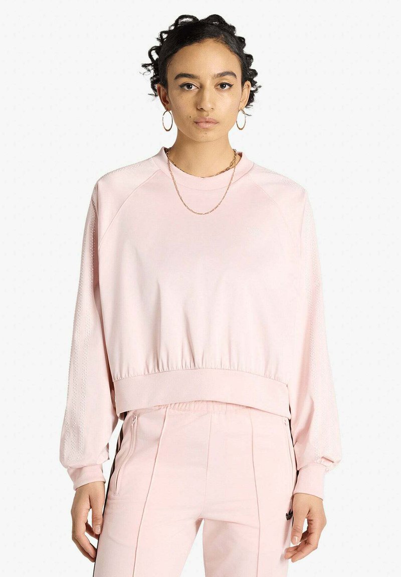 Femme portant un sweat-shirt à manches longues rose pâle et un pantalon assorti, debout face à l'avant sur un fond blanc uni.