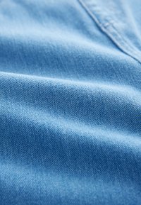 Tessuto di denim azzurro chiaro con una texture morbida, caratterizzato da motivi di tessitura visibili e lievi pieghe, indicativi di un materiale rilassato.