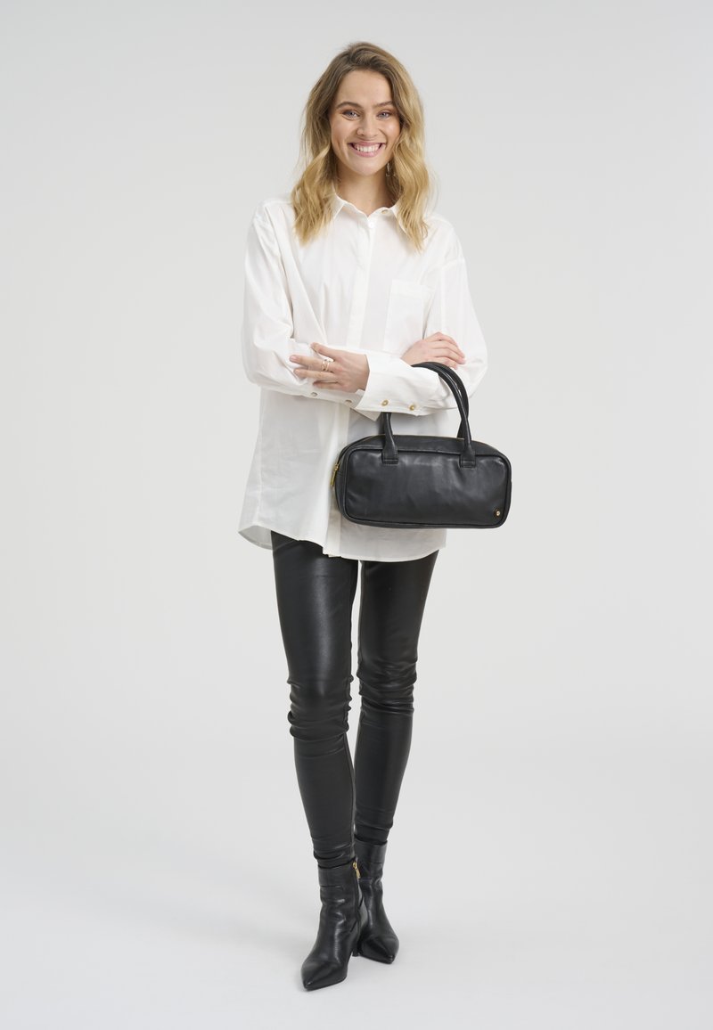 Jeune femme aux cheveux blonds souriante, portant une chemise blanche oversize, un pantalon en cuir noir et des bottes, tenant un sac à main noir devant un fond blanc.
