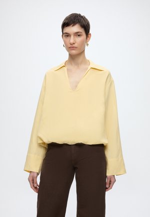 Άτομο με κοντά σκούρα μαλλιά που φοράει χρυσούς κρίκους στα αυτιά, μια φαρδιά ανοιχτό κίτρινη μπλούζα με μακριά μανίκια και σκούρο καφέ παντελόνι, στέκεται μπροστά σε ένα μονόχρωμο φόντο.