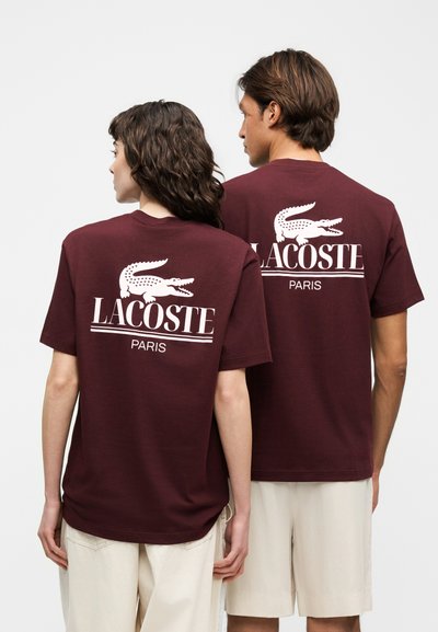 Двама души, облечени в еднакви бордови тениски Lacoste Paris с големи бели лого на крокодил на гърба, стоят един до друг.