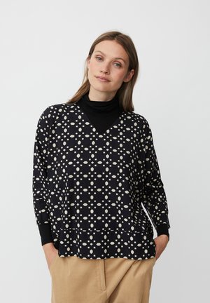 Top nero con scollatura a V, caratterizzato da un motivo geometrico bianco. Maniche lunghe con polsini neri. Indossato sopra un dolcevita nero e pantaloni beige.
