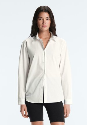 Žena s dlhými tmavými vlasmi, ktorá nosí nadrozmernú bielu košeľu na gombíky a čierne kraťasy po kolená, stojí pred jednoduchým bielym pozadím.