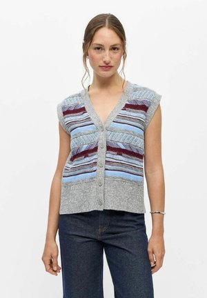 Femme à la peau claire et aux cheveux bruns portant un gilet en maille sans manches boutonné avec des rayures bleues et bordeaux et un jean bleu foncé.