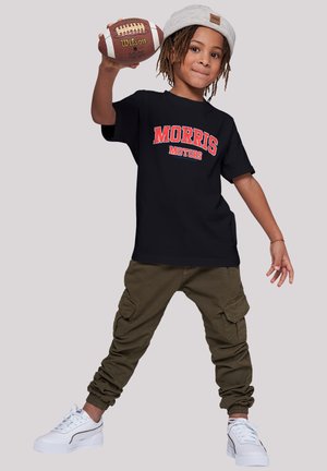 Black t-shirt with red "MORRIS MOTORS" graphic, olive cargo pants, and white sneakers; holding a brown football, wearing a gray beanie.
